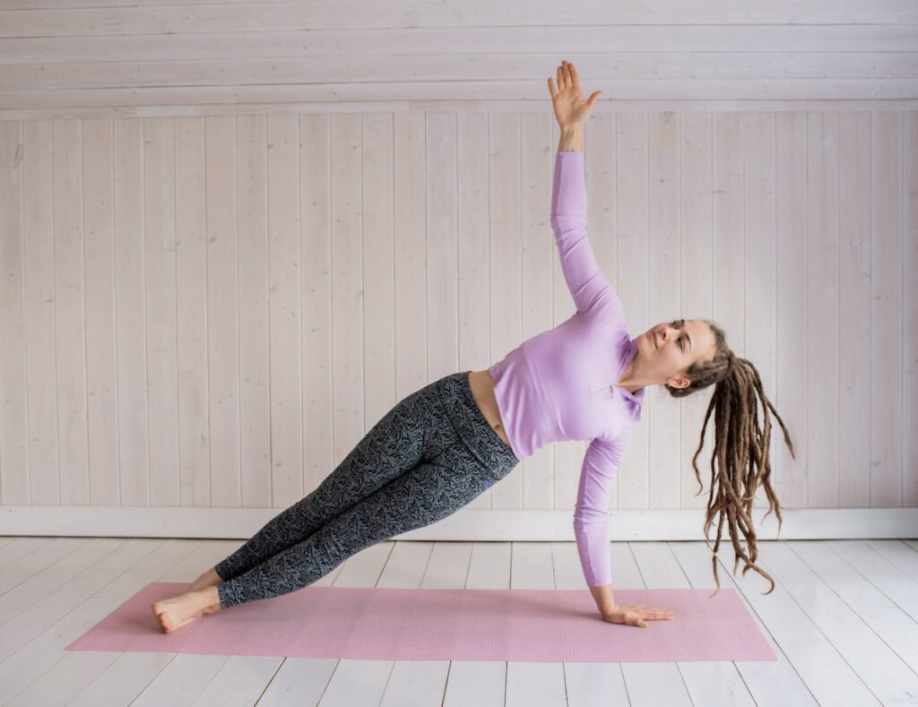 Woman in a side plank yoga pose on a pink mat in a bright indoor space.